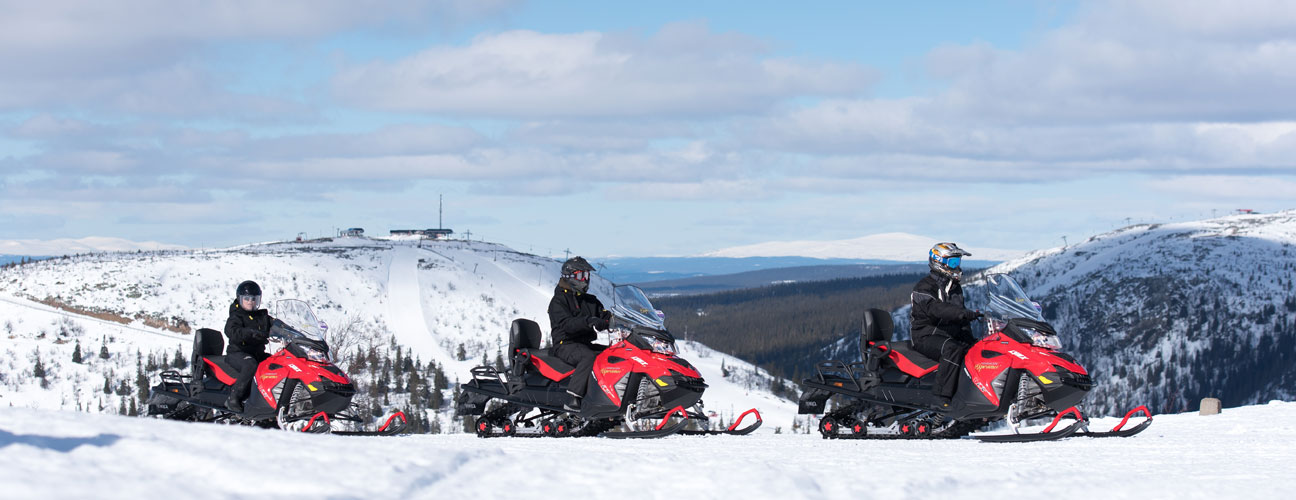 Tre röd snöskotrar med Vemdalen i bakgrunden, snörikt landskap med blå himmel.